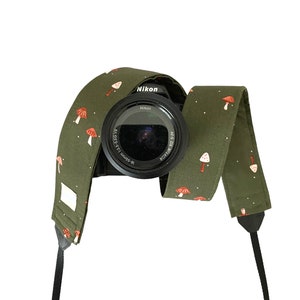 May include: A green camera strap with a white and red mushroom pattern. The strap is wrapped around a black camera with a Nikon lens.