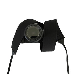 Peut inclure: Un sangle de caméra noire avec un appareil photo noir attaché. L'appareil photo a un objectif noir avec le mot "Nikon" imprimé dessus.
