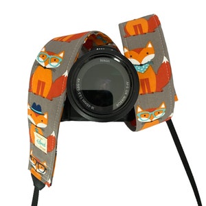 May include: A gray camera strap with a repeating pattern of orange foxes wearing glasses and blue bow ties. The strap is attached to a black camera lens with the word "Nikon" printed on it.