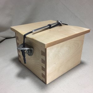 May include: A wooden box with a hinged lid and a metal latch. The box is made of light wood and has a natural finish. A black cord is attached to the lid and runs through a metal ring on the latch.