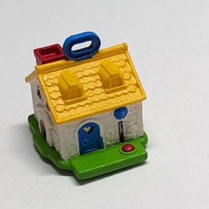 May include: A small white house with a yellow roof, blue door, and red chimney. The house has a blue loop on top and a red button on the front.