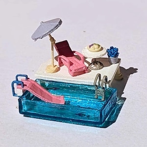 Puede incluir: Escena de piscina en miniatura con una piscina azul, un tobogán rosa y una mesa blanca con una tumbona rosa, una mesita con un plato y una planta azul en maceta. Una sombrilla lila proporciona sombra.