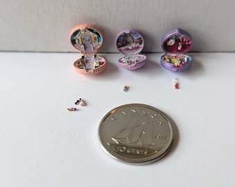 May include: Three miniature open compact cases in peach, light purple, and dark purple, each containing tiny interior scenes. Several minuscule figurines are scattered nearby, with a Canadian 10-cent coin providing a scale reference for size.