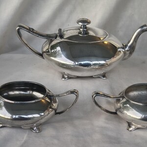 May include: A silver tea set including a teapot with a lid, a sugar bowl, and a creamer. The set is reflective and sits on a white surface. The teapot has a curved spout and handle. The sugar bowl and creamer have handles and sit on small feet.