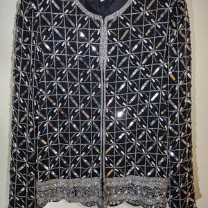 Könnte beinhalten: Eine schwarze Paillettenjacke mit einem geometrischen Muster in Silber und Weiß. Die Jacke hat einen Reißverschluss und einen gewellten Saum.