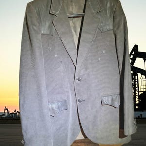 Può includere: Un blazer grigio in velluto a coste con inserti in pelle sulle spalle e sulle tasche. Il blazer ha una chiusura a due bottoni e un revers a lancia. La giacca è appesa a una gruccia nera.