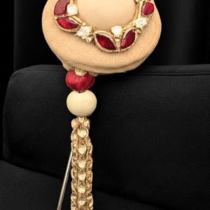 Peut inclure: Broche de style vintage avec un motif circulaire orné de pierres précieuses rouges et transparentes serties dans du métal doré. La broche est fixée à une base beige et ronde, avec une perle rouge et une perle crème, et une pampille dorée.