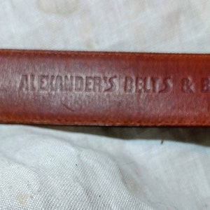May include: Close-up of a brown leather belt with the text "2987 ALEXANDER'S BELTS & BUCKLES" embossed on it.