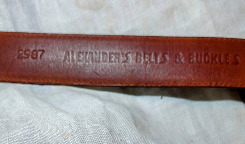 May include: Close-up of a brown leather belt with the text "2987 ALEXANDER'S BELTS & BUCKLES" embossed on it.
