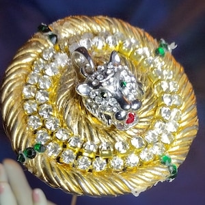 May include: A gold-tone brooch with a rhinestone-encrusted panther head. The panther has green and red accents and is surrounded by a circle of smaller rhinestones.