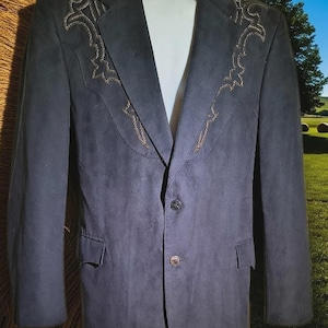 May include: A navy blue blazer with decorative gold-colored embroidery on the shoulders and lapels. The blazer has a single-button closure and two front pockets. The background shows a rural scene.