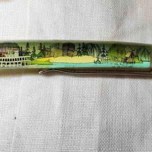 Op de afbeelding: Een pen met een doorzichtige schacht met een schilderachtige illustratie van een raderboot, bomen en gebouwen. De pen heeft een zwarte punt en een zilveren clip. Het kunstwerk is in tinten groen, geel en wit.