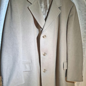 May include: A beige overcoat with a classic design. The coat features a notched lapel, three buttons, and two front pockets. The coat is hanging on a wooden hanger.