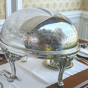 Puede incluir: Un chafing dish plateado con grabados florales ornamentados. La tapa abovedada y la base presentan detalles de volutas. El plato está sostenido por patas curvas decorativas y se encuentra sobre un mantel blanco.