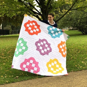 May include: A person holds up a white quilt with colorful squares in a checkered pattern. The quilt has a geometric design with rounded edges. The quilt is being held in a park setting with green grass and trees in the background.