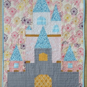May include: A quilted castle wall hanging featuring a blue and grey castle design. The castle has a pink and white interior and a yellow door. The background is a white fabric with a pink, purple and yellow floral pattern.
