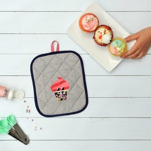 May include: A gray quilted hot pad with a blue trim and a pink and white cupcake applique. Three cupcakes with pink, white, and yellow frosting are on a white plate.