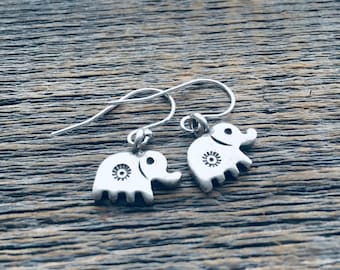 May include: A pair of silver elephant earrings with a simple design. The earrings are on a wooden surface.
