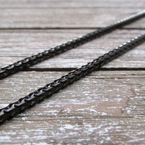 Puede incluir: Un collar de cadena negro hecho de peque&ntilde;os cuadrados de metal unidos. La cadena est&aacute; tendida sobre una superficie de madera.