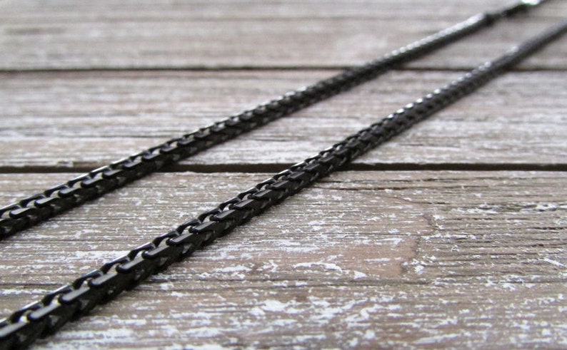 Puede incluir: Un collar de cadena negro hecho de peque&ntilde;os cuadrados de metal unidos. La cadena est&aacute; tendida sobre una superficie de madera.