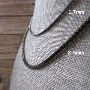 Puede incluir: Dos collares de cadena negra sobre un fondo de tela gris. La cadena superior tiene 1,7 mm de grosor y la cadena inferior tiene 2,5 mm de grosor.