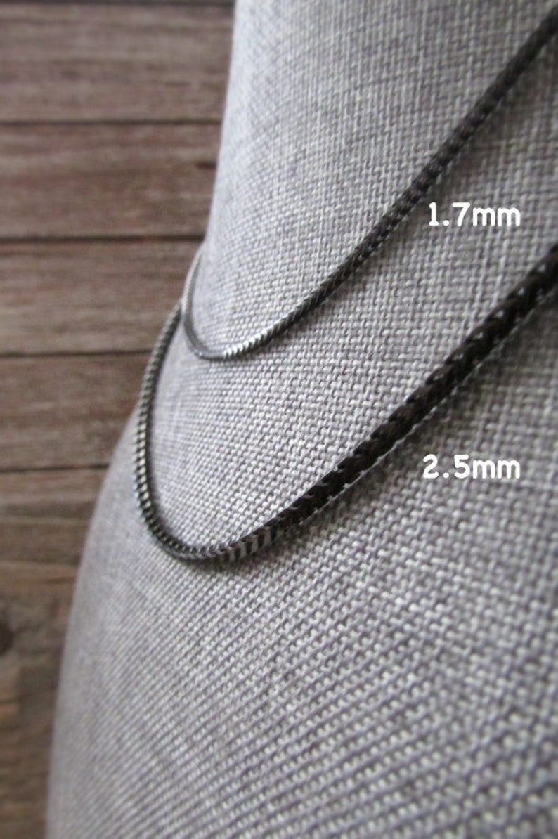 Puede incluir: Dos collares de cadena negra sobre un fondo de tela gris. La cadena superior tiene 1,7 mm de grosor y la cadena inferior tiene 2,5 mm de grosor.
