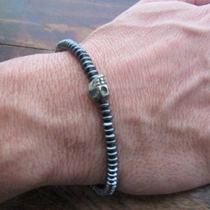 May include: A black hematite bracelet with a silver skull charm. The bracelet is made of small, round beads and is worn on a person's wrist.