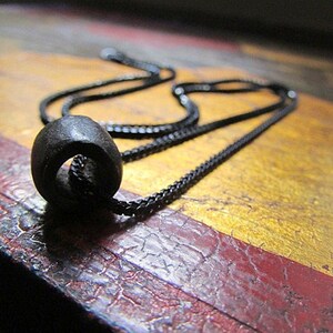 Puede incluir: Un collar de cadena negra con un colgante redondo negro. El colgante est&aacute; hecho de un material liso y oscuro.