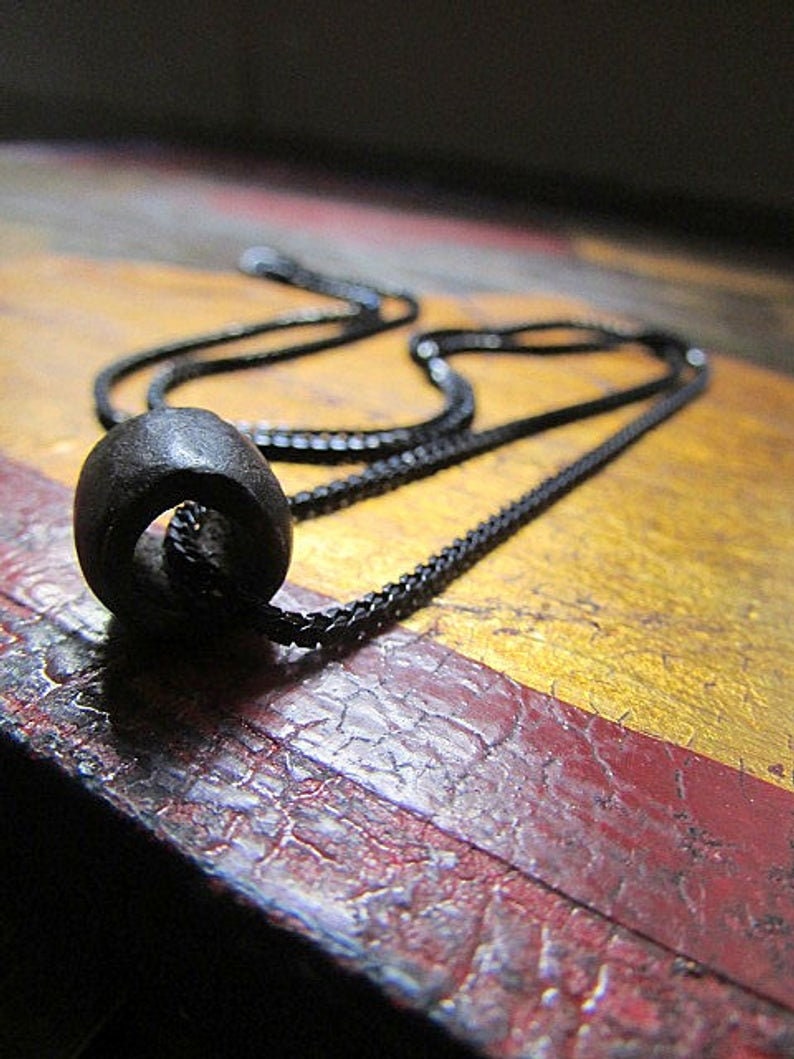 Puede incluir: Un collar de cadena negra con un colgante redondo negro. El colgante est&aacute; hecho de un material liso y oscuro.