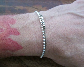Puede incluir: Un brazalete de cadena de plata con un cordón verde. El brazalete está en la muñeca de una persona.