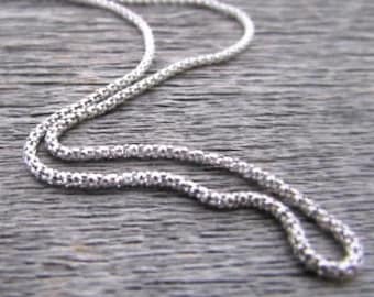 Könnte beinhalten: Eine silberne Kettenkette mit einem zarten, strukturierten Design. Die Kette besteht aus kleinen, runden Gliedern, die eng miteinander verwoben sind.