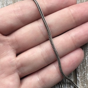 May include: A silver chain necklace with a delicate, braided design. The chain is shown lying flat on a hand.