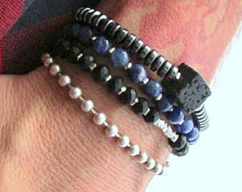 Puede incluir: Un conjunto de pulseras con cuentas negras, plateadas y azules. La pulsera superior tiene una cuenta de lava negra.