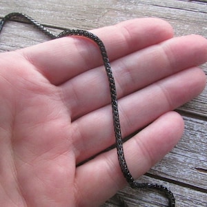 Puede incluir: Un collar de cadena negro con un dise&ntilde;o trenzado. La cadena est&aacute; sobre la mano de una persona.