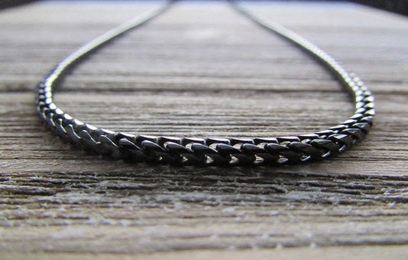 Puede incluir: Un collar de cadena negro hecho de eslabones de metal. La cadena est&aacute; sobre una superficie de madera.