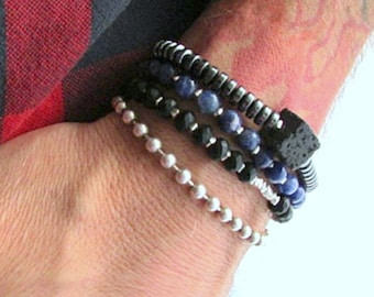 Puede incluir: Un conjunto de pulseras con cuentas negras, azules y plateadas. Las pulseras están superpuestas en la muñeca de una persona.