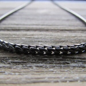 Puede incluir: Un collar de cadena negro hecho de eslabones de metal. La cadena est&aacute; sobre una superficie de madera.