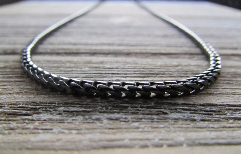 Puede incluir: Un collar de cadena negro hecho de eslabones de metal. La cadena est&aacute; sobre una superficie de madera.