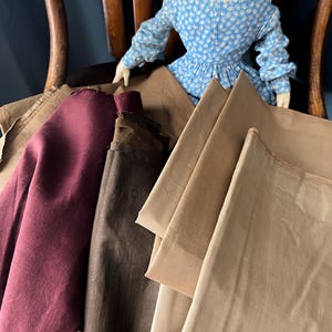 May include: Assortment of fabric swatches in various shades of brown, burgundy, and tan. A doll in a blue floral dress sits in the background. The fabrics are arranged on a wooden chair, showcasing their textures and colors.