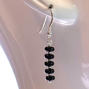 May include: A single dangle earring with a silver hook and a series of five black, oval-shaped beads. The beads are strung together with silver links, creating a simple yet elegant design. The earring is shown against a blurred background.