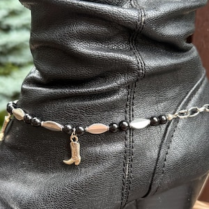 May include: A black leather boot with a silver and black beaded boot chain. The chain features silver oval beads, black round beads, and a gold-colored cowboy boot charm.