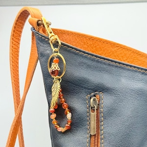 May include: A gold-colored charm with a beaded teardrop design and a gold-colored feather attached to a black and orange leather bag. The charm has a gold-colored ring with a decorative element and a red bead.