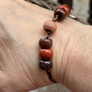 May include: A beaded bracelet featuring a series of round, earthy-toned beads in shades of brown, orange, and speckled black. The beads are strung on a black and orange cord, with a silver clasp.