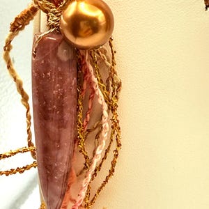 May include: Close-up of a handmade pendant featuring a polished, elongated, reddish-brown stone, a large gold-colored bead, and decorative strands of gold and pink thread. The pendant is suspended from a gold-colored metal finding.