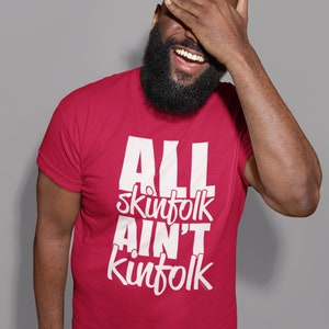 May include: A crimson t-shirt with the white text "ALL skinfolk AIN'T kinfolk". The person wearing the shirt is smiling and covering their face with their hand. The background is a plain grey.
