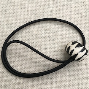 May include: A black hair tie with a decorative bead. The bead is cylindrical, with a cream-colored base and a black zebra stripe pattern. The hair tie is made of a stretchy black material.