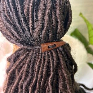 May include: Close-up of dark brown dreadlocks styled with a wooden hair accessory. The accessory is a small, rectangular piece of wood with a hole for the elastic band. The dreadlocks are neatly arranged.