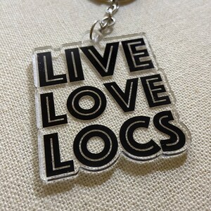 Puede incluir: Llavero de acrílico transparente con las palabras "LIVE LOVE LOCS" en letras negras en negrita. El llavero tiene un anillo y una cadena de metal plateado para sujetarlo a llaves o a un bolso. El fondo es de tela beige claro.
