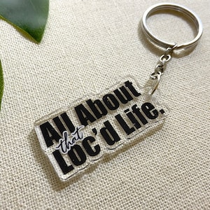 Puede incluir: Llavero acrílico transparente con el texto "All About that Loc'd Life" en negro. El llavero tiene un llavero y una cadena plateados. El fondo es una tela neutra y texturizada.
