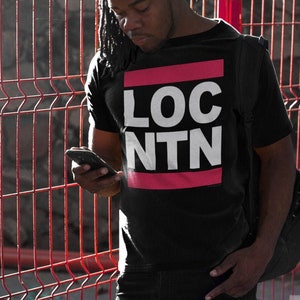 May include: Black t-shirt with the text "LOC NTN" in white letters, framed by a pink rectangle. The person is wearing black jeans and a black backpack. The background features a red metal fence.